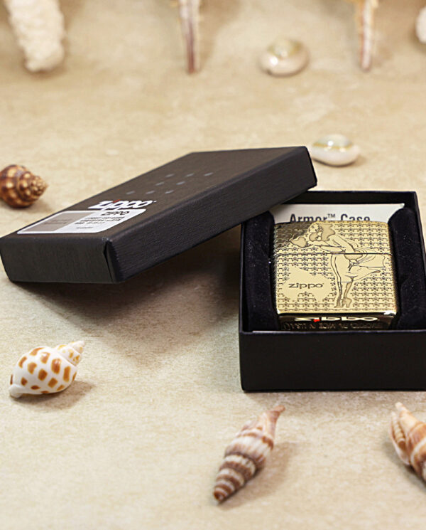 Gold engraved Zippo lighter in an open black presentation box on a sandy surface with seashells around it.