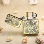 Gold engraved Zippo-style lighter open on a sandy beach with scattered seashells nearby, intricate patterns visible on the metal.
