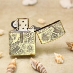 Gold ornate lighter on its side with lid open, placed on sand among seashells; decorative engravings visible on the metal surface.