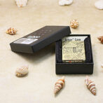 Ornate metal lighter in an open black box labeled Armor Case, surrounded by small seashells on a sandy surface.