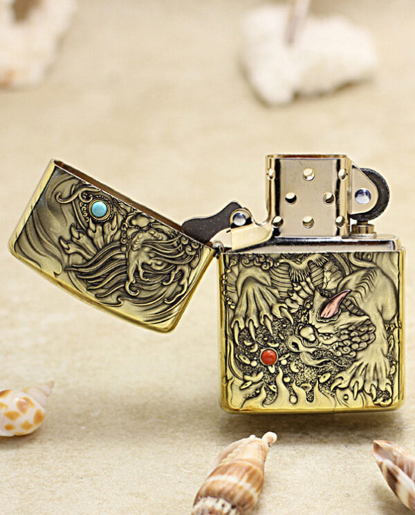 Gold engraved dragon lighter open to reveal ornate designs, resting on sandy beach with seashells nearby