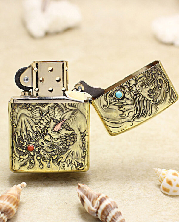 Gold-tone lighter with dragon relief engraving on a sandy beach, lid open among seashells