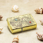 Gold ornate lighter with a dragon engraving and blue/red gemstone accents, set on sand with seashells nearby.