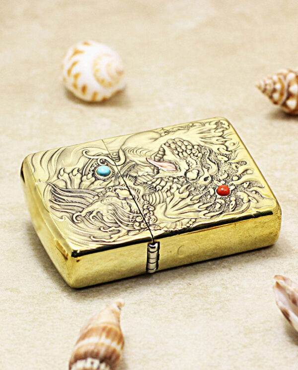 Gold ornate lighter with a dragon engraving and blue/red gemstone accents, set on sand with seashells nearby.