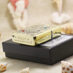 Gold engraved Zippo lighter resting on a black box, with seashells in the foreground.