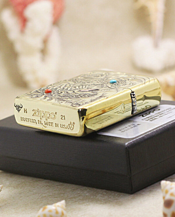 Gold engraved Zippo lighter resting on a black box, with seashells in the foreground.