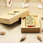 Brass engraved dragon lighter in an open wooden gift box on a sandy beach with seashells nearby.