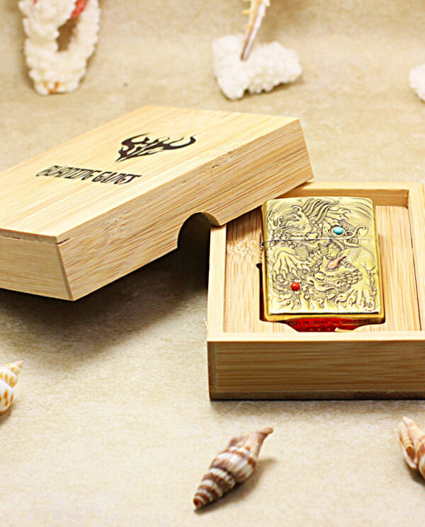Brass engraved dragon lighter in an open wooden gift box on a sandy beach with seashells nearby.