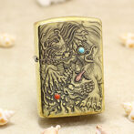 Gold-tone Zippo lighter with an intricate dragon engraving, featuring a turquoise eye and red orb, on a sandy surface with seashells nearby.