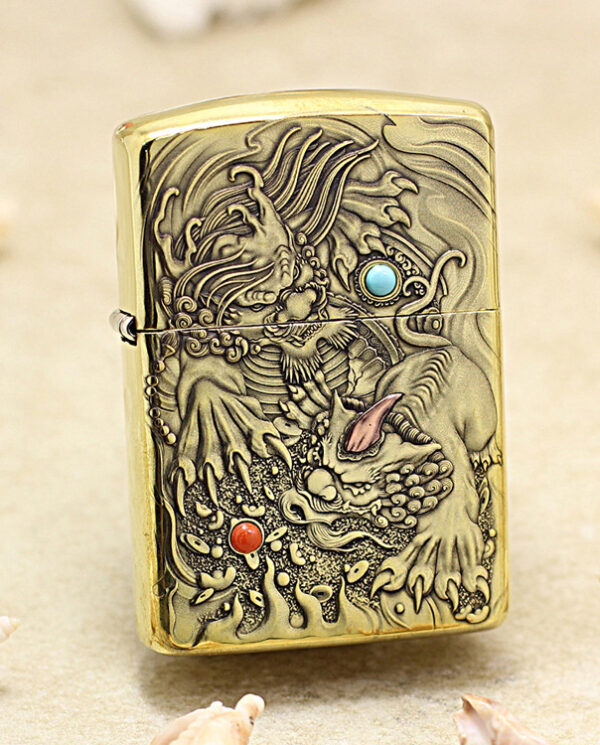 Gold-tone Zippo lighter with an intricate dragon engraving, featuring a turquoise eye and red orb, on a sandy surface with seashells nearby.
