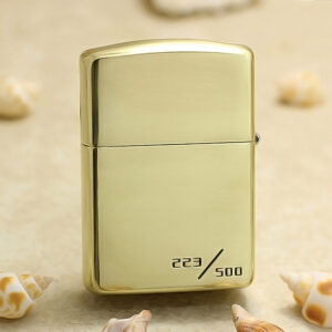 Gold Zippo-style lighter with numbered edition 223/500 on a sandy background with small seashells nearby