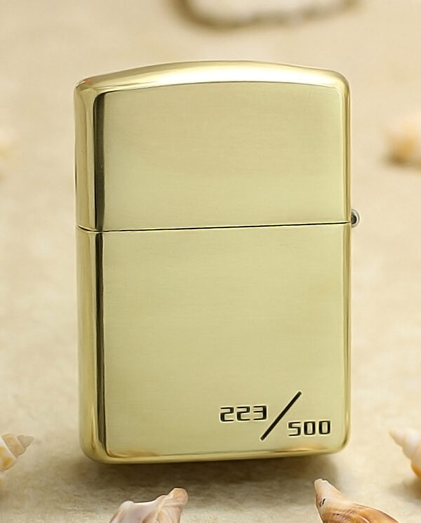 KZ253-01 Gold Zippo-style lighter with numbered edition 223/500 on a sandy background with small seashells nearby