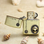 Gold Zippo-style lighter open on a sandy beach, featuring an alien head design on the wheel amid seashells nearby.