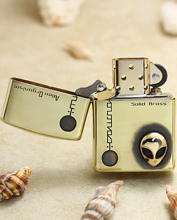 KZ253-02 Gold Zippo-style lighter open on a sandy beach, featuring an alien head design on the wheel amid seashells nearby.