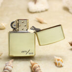 Gold Zippo lighter with open lid resting on sandy beach, surrounded by seashells; engraved '223/500' on its bottom.
