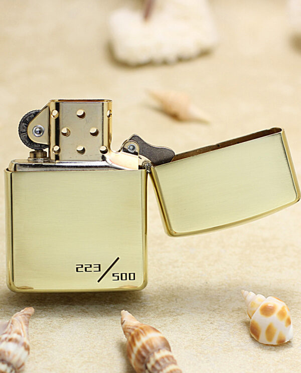 KZ253-03 Gold Zippo lighter with open lid resting on sandy beach, surrounded by seashells; engraved '223/500' on its bottom.