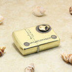 Gold Zippo-style lighter lying on sand with seashells scattered around it, decorative and beachy.