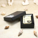 A small metal lighter in an open black box on a sandy surface with seashells scattered around.