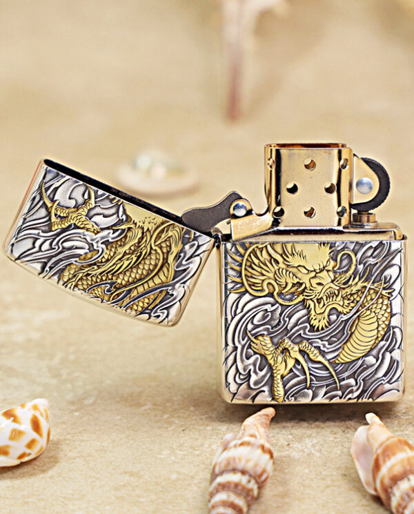 KZ282-02 Metallic lighter with gold dragon engraving, open lid, resting on a sandy surface with seashells nearby.