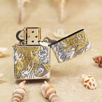 Decorative metal lighter with intricate gold and silver floral engraving, open lid, on sandy beach with seashells nearby.