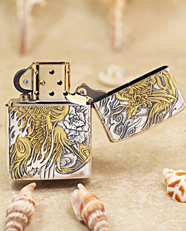 KZ282-03 Decorative metal lighter with intricate gold and silver floral engraving, open lid, on sandy beach with seashells nearby.