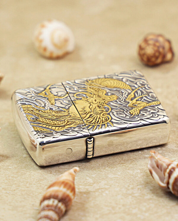 KZ282-04 Metal lighter with gold dragon engraving on a silver body, resting on sand among seashells.
