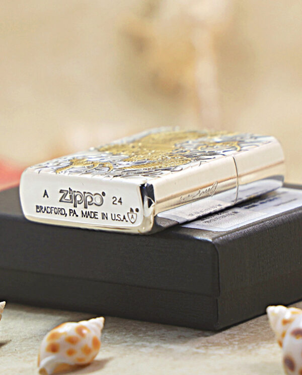 KZ282-05 Shiny Zippo lighter with an ornate engraved pattern resting on a black box, with small seashells scattered on sand nearby.