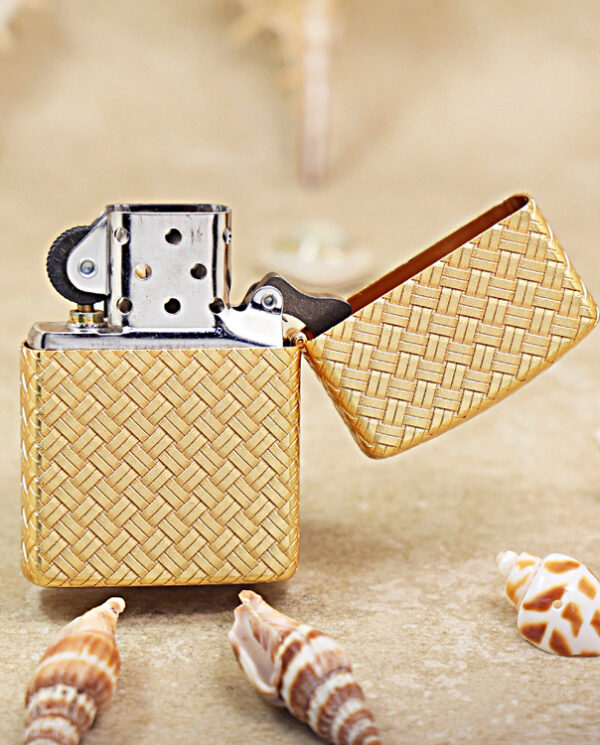 KZ284-03 Gold woven metal lighter with open lid resting on a sandy surface, surrounded by seashells (informative, foreground focus).