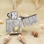 Open, ornately engraved metal lighter with floral scrollwork and a gold skull emblem, resting on sand among seashells.