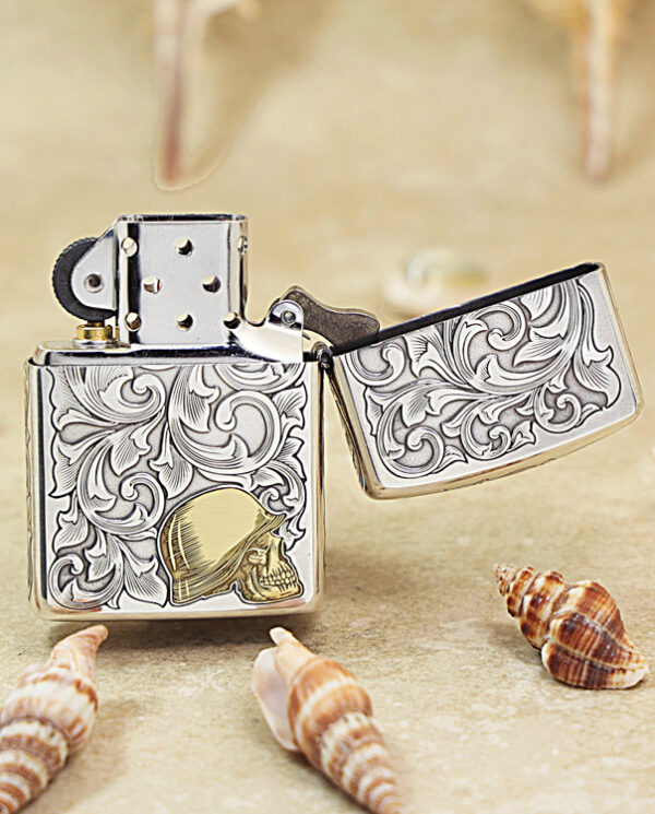 KZ286-03 Open, ornately engraved metal lighter with floral scrollwork and a gold skull emblem, resting on sand among seashells.