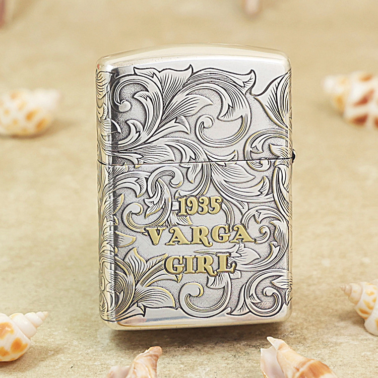 Metal lighter with intricate scroll engraving and gold lettering reading '1935 VARGA GIRL' on a sandy background.