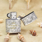 Engraved metal lighter with swirling floral patterns and the text '1935 Varga Girl' on a sandy beach with seashells nearby.