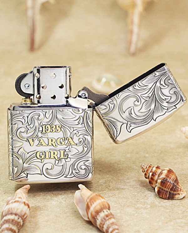 KZ287-03 Engraved metal lighter with swirling floral patterns and the text '1935 Varga Girl' on a sandy beach with seashells nearby.