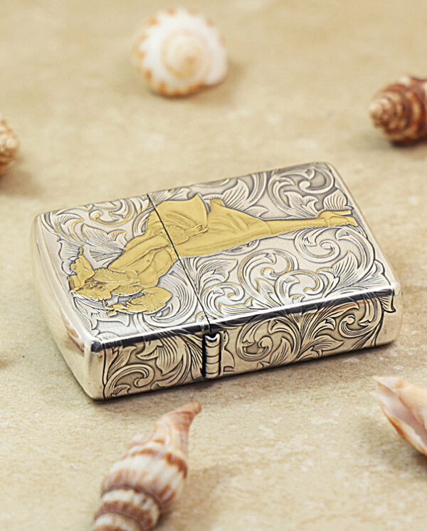 KZ287-04 Ornate silver cigarette lighter with swirling engravings and a gold silhouette of a reclining figure, placed on sand among seashells.