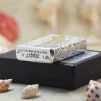 Close-up of a decorative Zippo lighter with an intricate silver pattern resting on a black box, with seashells in the foreground.