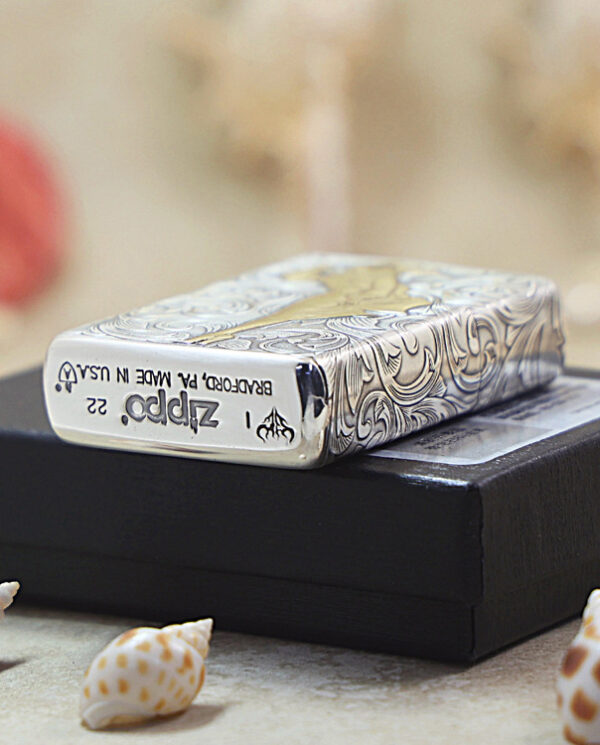 KZ287-05 Close-up of a decorative Zippo lighter with an intricate silver pattern resting on a black box, with seashells in the foreground.