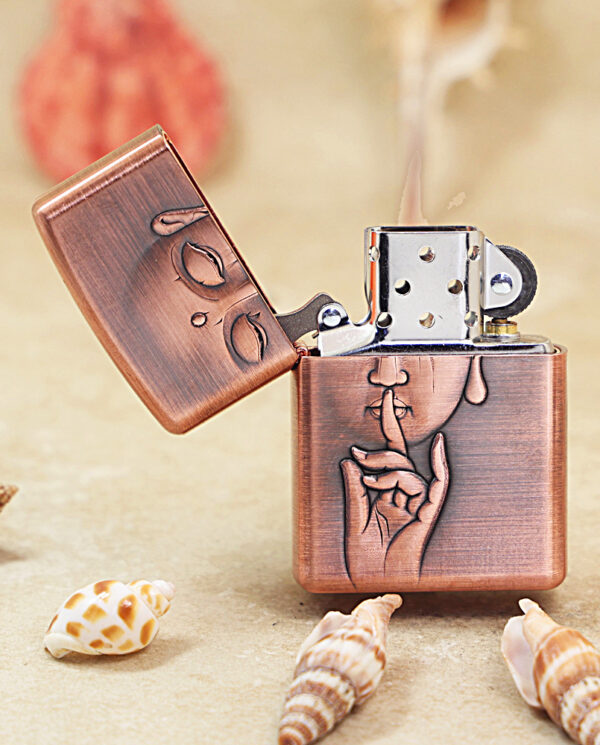 KZ288-02 Copper Zippo lighter with hand‑to‑mouth 'shh' engraving, lid open, on a sandy beach with seashells nearby