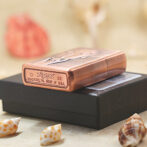 Copper Zippo lighter resting on a black box, with seashells in the foreground and a blurred background. ASText: a product photo showing a copper Zippo lighter on a matte black box with small seashells around.