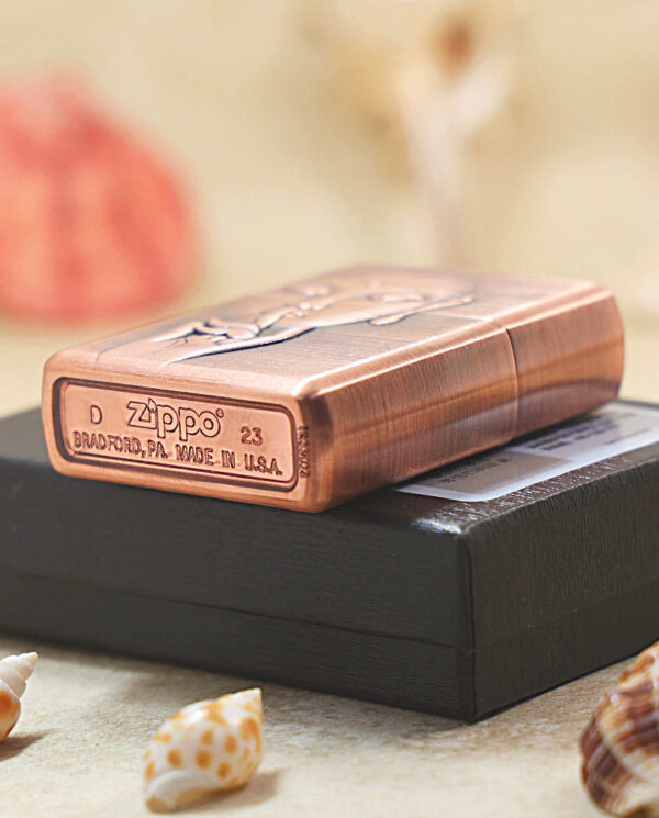 KZ288-04 Copper Zippo lighter resting on a black box, with seashells in the foreground and a blurred background. ASText: a product photo showing a copper Zippo lighter on a matte black box with small seashells around.