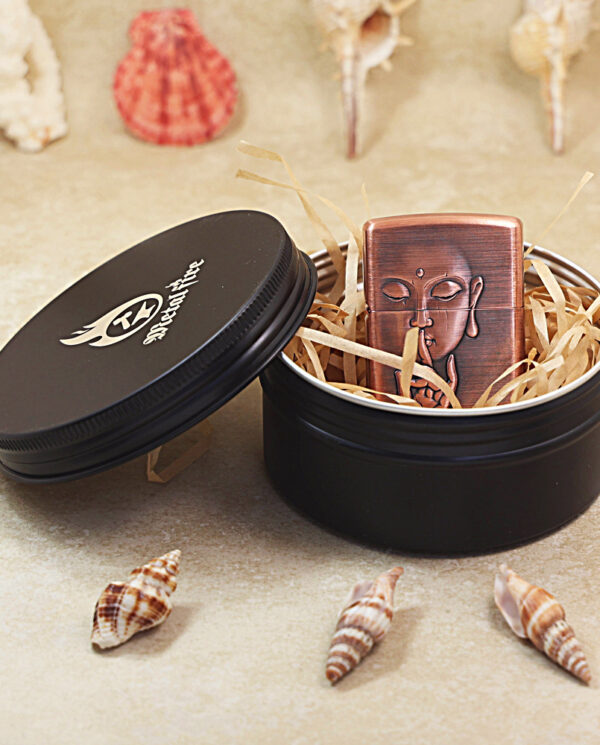 KZ288-05 Open black circular tin revealing a copper Buddha-design lighter nestled in straw filling, with seashells in the background.