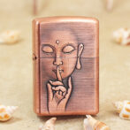 Copper-colored lighter engraved with a calm face making a shushing gesture (finger to lips); shells in a sandy background.