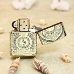 Ornate vintage metal lighter with open lid on sandy beach, surrounded by seashells.