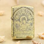 Gold Zippo-style lighter with an intricate Buddhist deity engraving and floral patterns on a sandy surface.