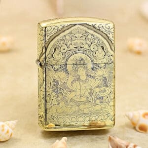 Gold Zippo-style lighter with an intricate Buddhist deity engraving and floral patterns on a sandy surface.