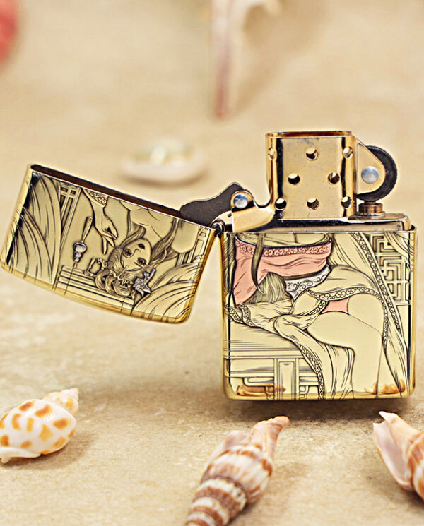 KZ426-02 Gold engraved lighter with ornate illustration, open lid, resting on a sandy beach with shells nearby.