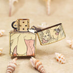 Gold metal lighter with colorful pin-up girl artwork on its sides, open lid, resting on a sandy surface with seashells nearby.
