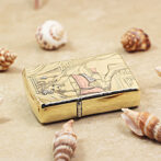 Gold-toned metal lighter with an engraved image of a woman in traditional dress, resting on sand among seashells.