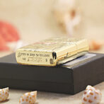 Gold Zippo lighter with an engraved design rests on a black box, with small seashells in the foreground.