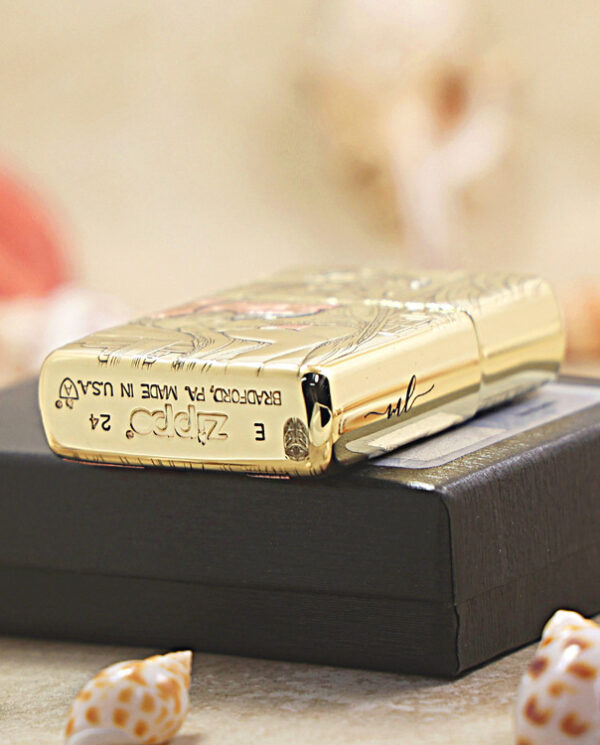 KZ426-05 Gold Zippo lighter with an engraved design rests on a black box, with small seashells in the foreground.