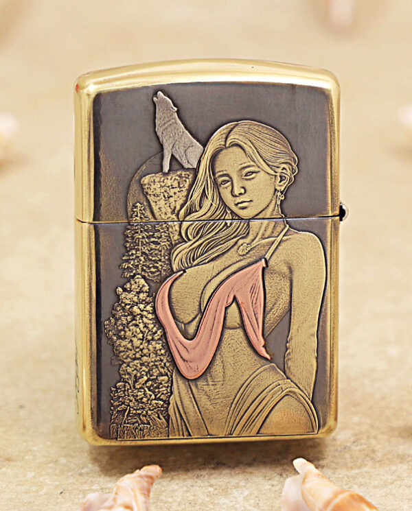 KZ452-01 Brass Zippo-style lighter with an engraved panel showing a woman in a pink bikini top beside a forest scene, set on a sandy background with seashells nearby.